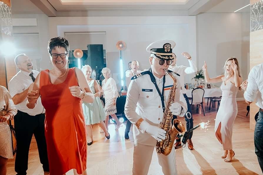 Saksofonista w białym mundurze marynarskim gra na saksofonie podczas imprezy, w tle tańczący goście i dekoracje.