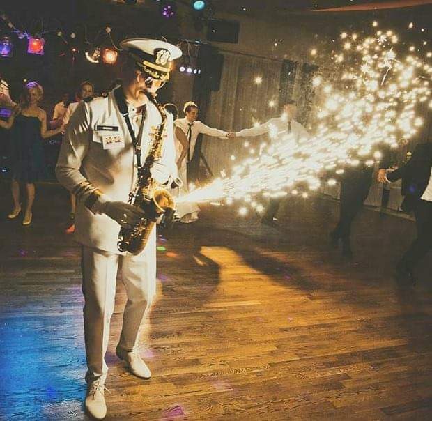 Saksofonista w białym mundurze marynarskim gra na saksofonie, z którego wydobywają się iskry, podczas gdy w tle tańczą goście na parkiecie z drewnianą podłogą.