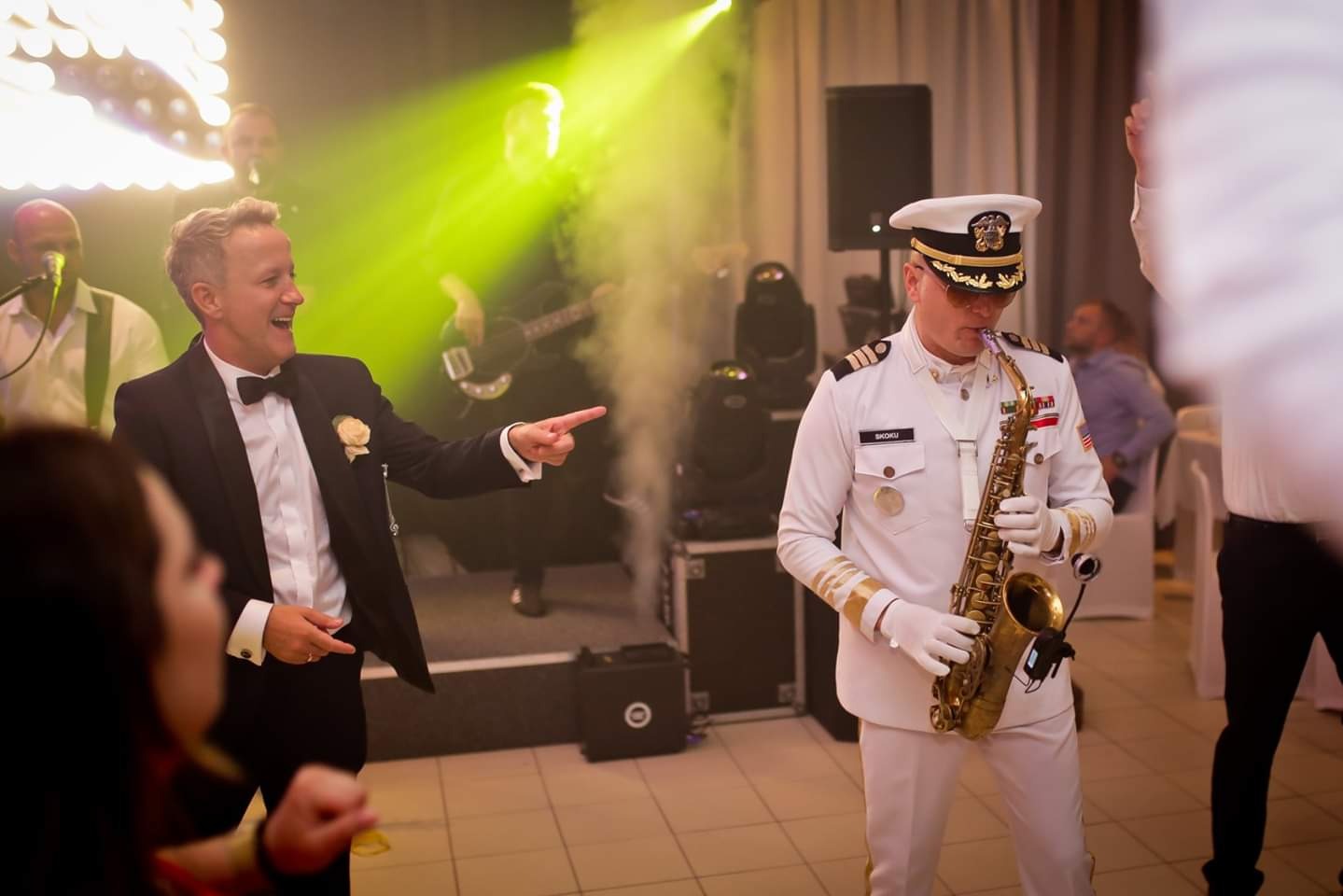 Saksofonista w białym mundurze marynarki wojennej gra na saksofonie podczas występu zespołu na imprezie, mężczyzna w smokingu wskazuje na niego z uśmiechem.