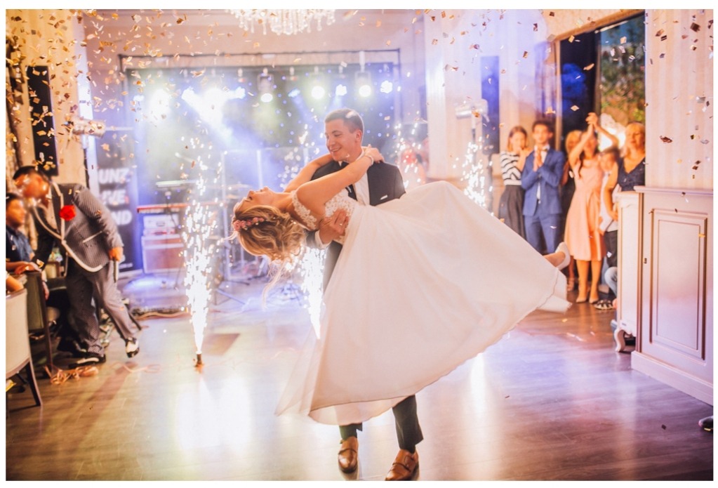 Para młoda w tańcu, otoczona fontannami iskier i opadającym konfetti, w tle zespół muzyczny i goście weselni.