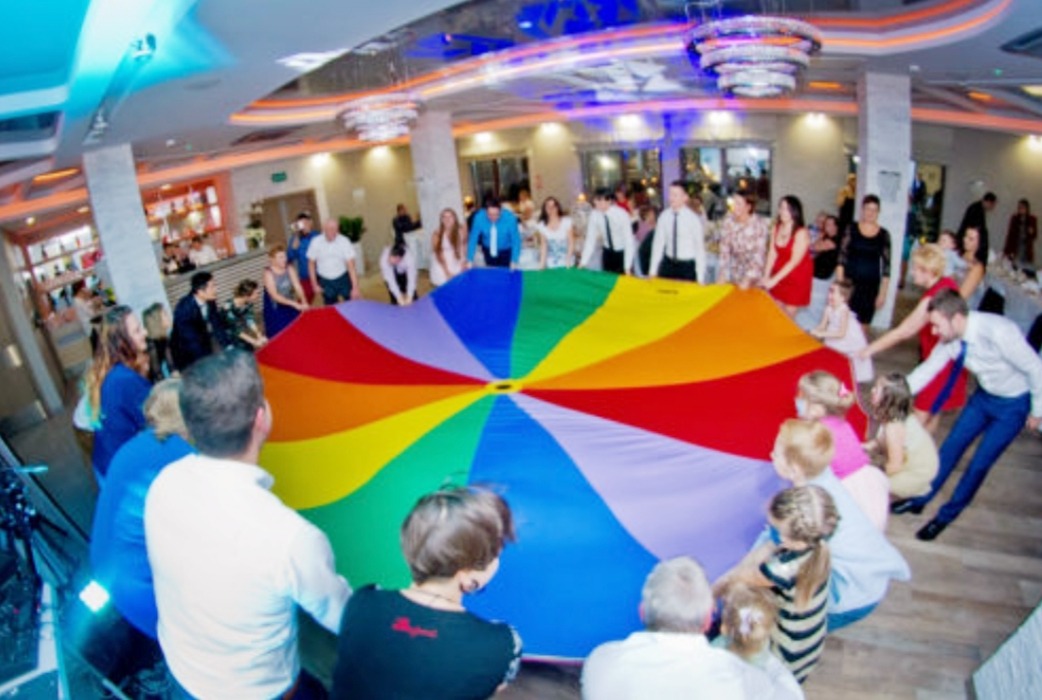 Grupa dorosłych i dzieci trzymająca kolorową chustę animacyjną w sali bankietowej z dekoracyjnymi żyrandolami, podczas zabawy integracyjnej.