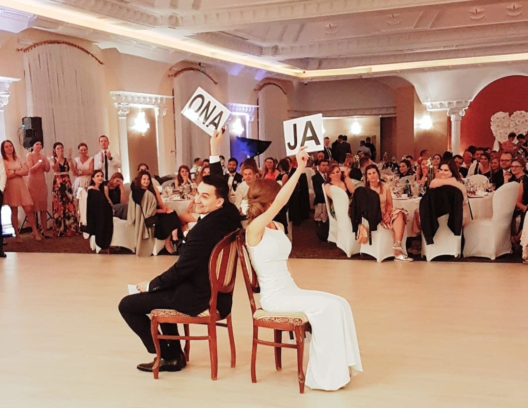 Para młoda siedzi tyłem do siebie na krzesłach podczas zabawy weselnej, trzymając kartki z napisami 'ONA' i 'JA', goście weselni w tle.