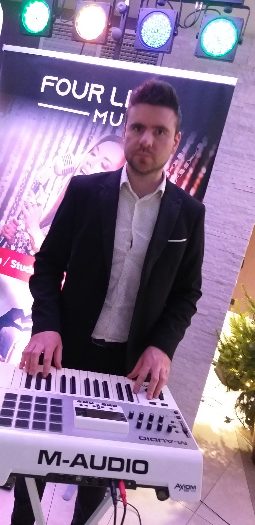 Mężczyzna w czarnej marynarce gra na białym keyboardzie M-Audio Axiom przed banerem z wizerunkiem wokalistki i napisem 'FOUR LINES MU'. W tle oświetlenie sceniczne i fragment dekoracji świątecznej.