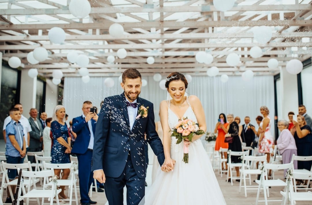 Uśmiechnięta para młoda w Krakowie, w eleganckich strojach, opuszcza ceremonię ślubną pod baldachimem białych lampionów, obsypywana konfetti przez gości.