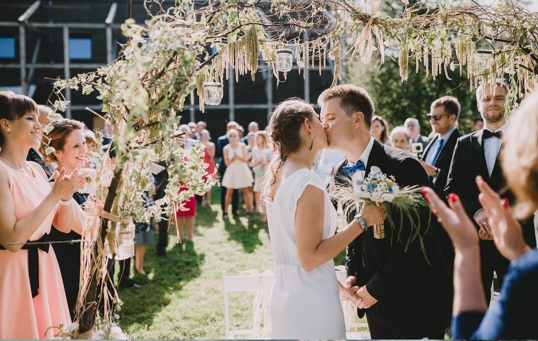 Pocałunek pary młodej pod rustykalnym łukiem z gałęzi, zdobionym wiszącymi słoikami i suszem, w tle goście weselni klaszczący w dłonie.