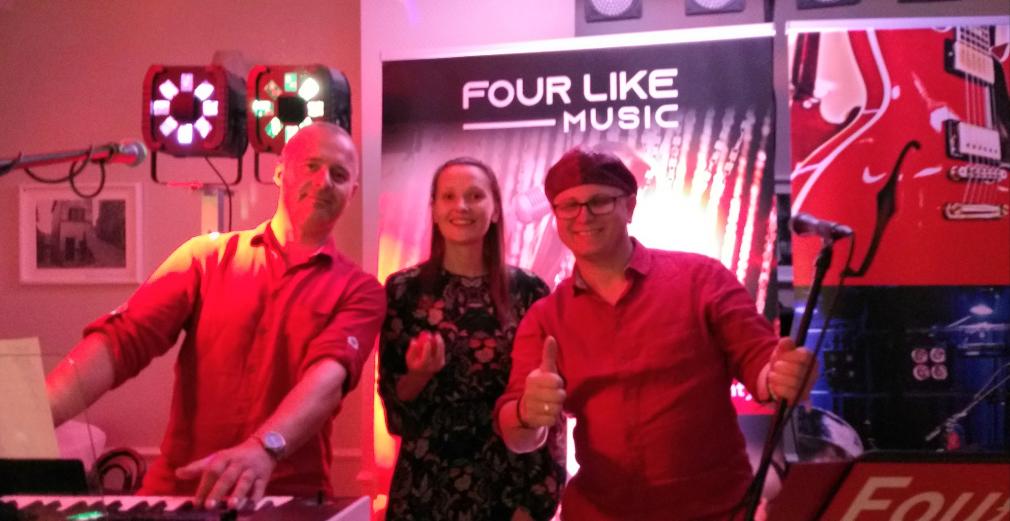 Trzyosobowy zespół pozuje przed banerem z nazwą 'Four Like Music', mężczyzna w czerwonej koszuli gra na keyboardzie, drugi mężczyzna w czerwonej koszuli z uniesionym kciukiem trzyma mikrofon,...