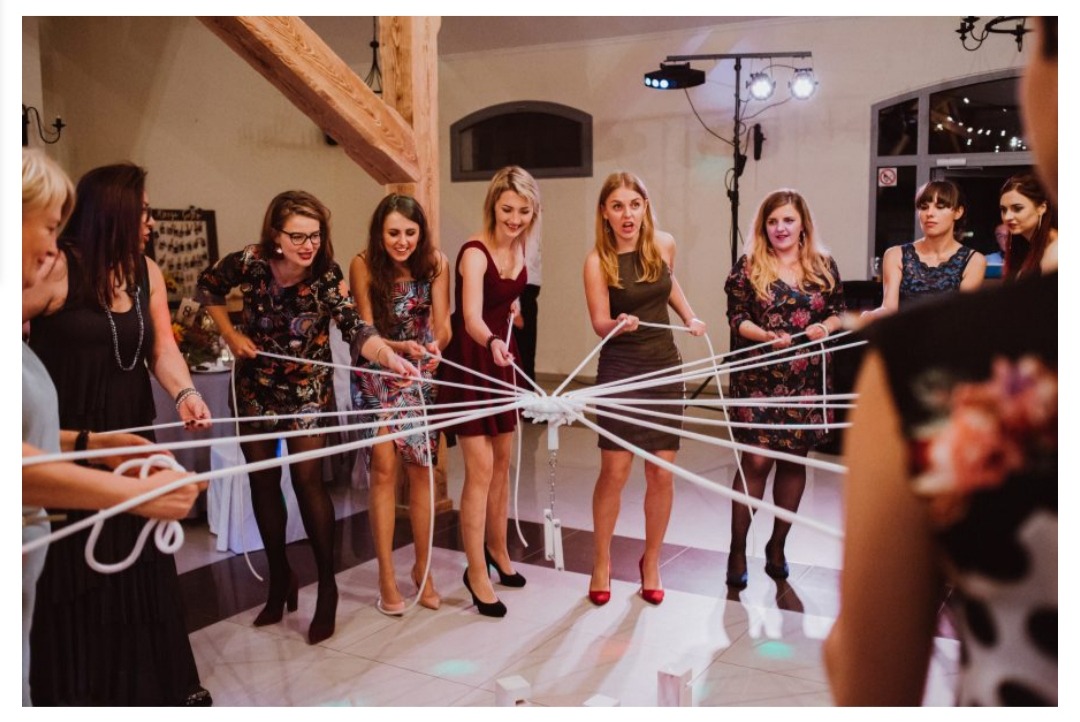 Grupa kobiet w sukienkach uczestniczy w zabawie weselnej z białymi sznurkami, tworzącymi pajęczynę wokół centralnego punktu, w tle drewniana konstrukcja i oświetlenie sceniczne.