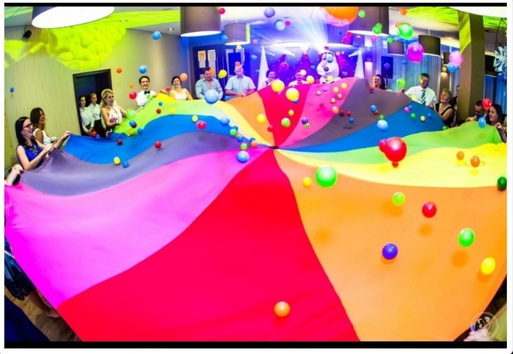 Grupa osób trzymająca dużą, kolorową chustę animacyjną z unoszącymi się w powietrzu kolorowymi piłeczkami podczas zabawy w sali z dekoracjami.