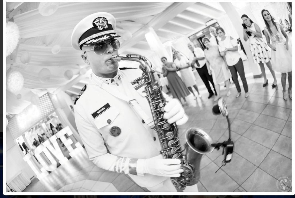 Czarno-białe zdjęcie saksofonisty w białym mundurze galowym, grającego na saksofonie podczas uroczystości, z dekoracjami i publicznością w tle. Ujęcie z dołu, perspektywa rybiego oka.