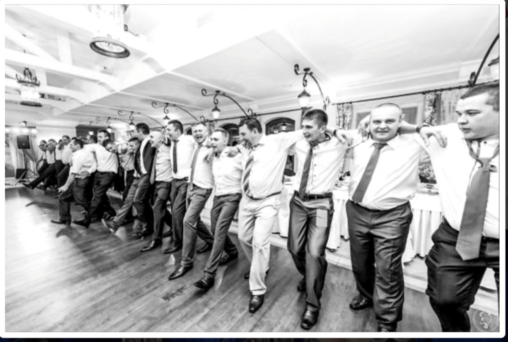 Czarno-białe zdjęcie grupy mężczyzn w koszulach i krawatach, tańczących w rzędzie z rękami na ramionach, na drewnianej podłodze w oświetlonym pomieszczeniu z dekoracyjnymi lampami.