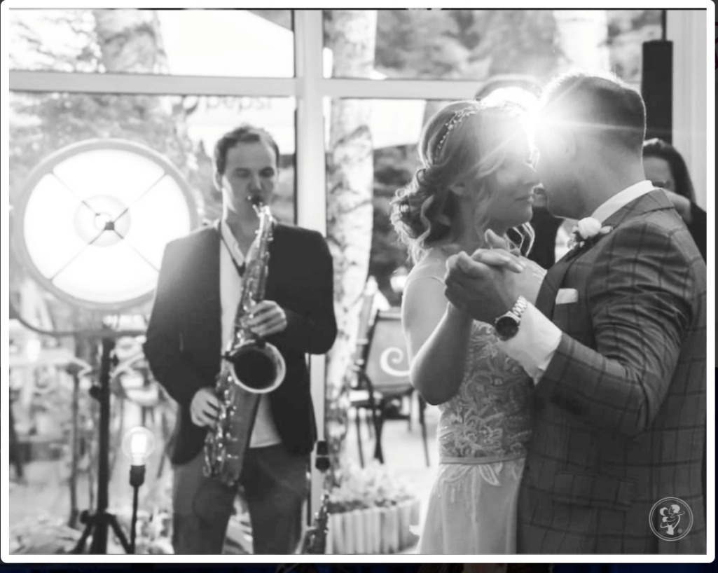 Czarno-białe zdjęcie pary młodej w tańcu podczas wesela, z saksofonistą w tle i dekoracyjnym oświetleniem.