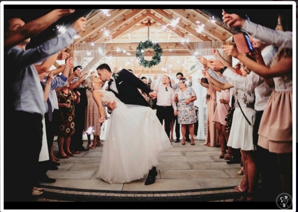 Nowożeńcy w trakcie pierwszego tańca, otoczeni gośćmi z odpalonymi zimnymi ogniami, dekoracja sali z drewnianą konstrukcją i zielonym wieńcem.