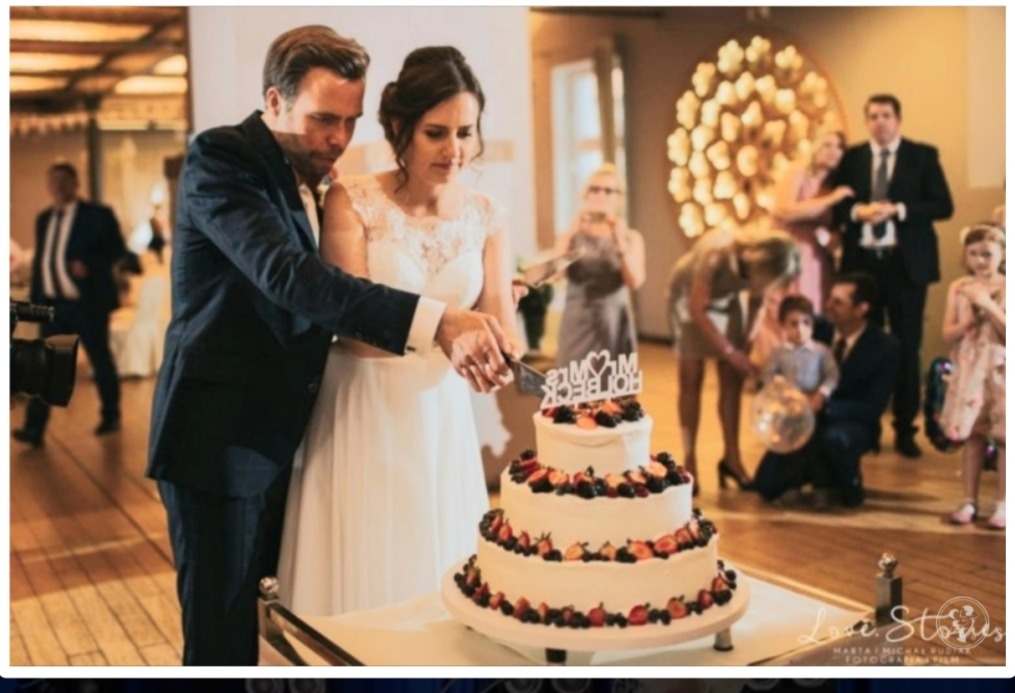 Nowożeńcy kroją tort weselny, trzy piętra dekorowane truskawkami i jeżynami, z topperem 'Mr&Mrs Holbeck', goście w tle.