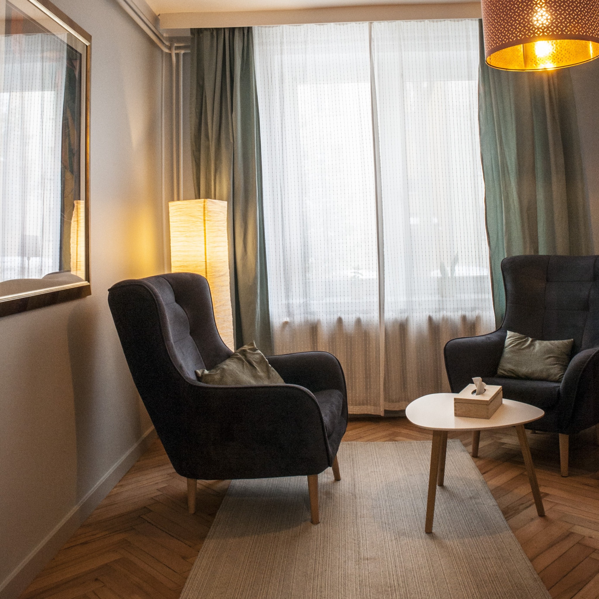 Ustawienie pokoju: Dwa ciemne fotele z poduszkami, mały okrągły stolik z pudełkiem chusteczek, dywan w paski, drewniana podłoga w jodełkę, lampa podłogowa i wisząca lampa z ażurowym abażurem, okno...