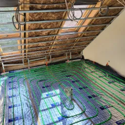 Instalacja ogrzewania podłogowego z zielonych rurek na folii z nadrukiem, widok z dołu na sufit z izolacją i oknem dachowym.