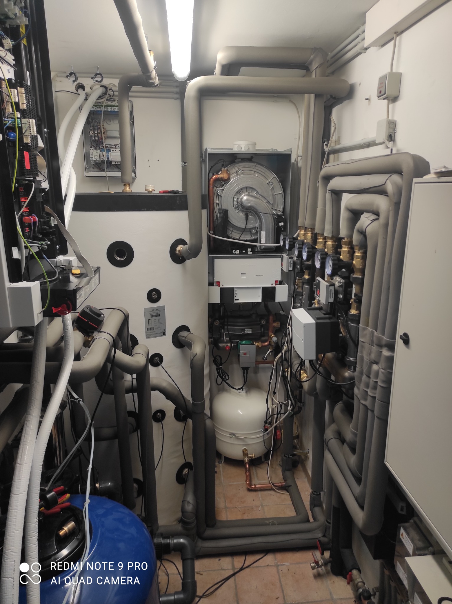 Węzeł cieplny w kotłowni, widoczne liczne rury w izolacji, zawory, manometry, zbiornik wyrównawczy, piec CO z wentylatorem, oraz rozdzielnica elektryczna na ścianie.