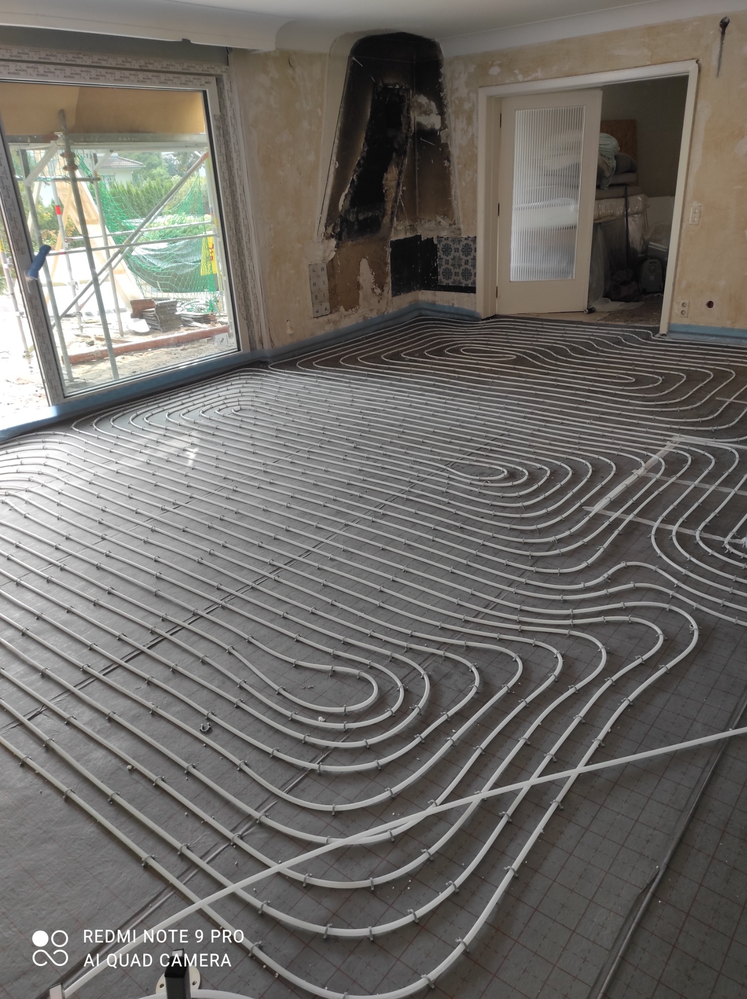 Białe rurki ogrzewania podłogowego ułożone spiralnie na szarej macie w trakcie instalacji w pokoju z oknem balkonowym i widocznymi śladami prac remontowych na ścianach.