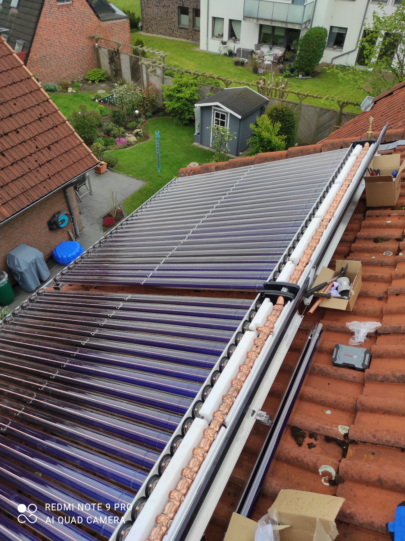 Montaż kolektorów słonecznych na dachu z czerwonej dachówki, widoczne narzędzia w kartonach, tło stanowią budynki i ogród