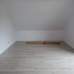 Poddasze po montażu jasnych paneli podłogowych, widoczne kartony z panelami w centralnej części pomieszczenia, ściany pomalowane na biało, brązowy żyrandol na suficie.