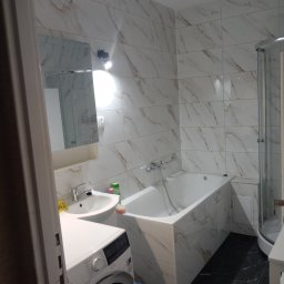 Ogólnobudowlane - Wykończona łazienka z wanną obudowaną płytkami imitującymi marmur, kabiną prysznicową i pralką. Umywalka z lustrem i czarna podłoga.