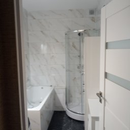 Ogólnobudowlane - Nowoczesna łazienka po remoncie: białe marmurowe płytki, kabina prysznicowa z zaokrąglonym szkłem, wanna obudowana płytkami i czarna podłoga. Widok zza uchylonych drzwi.