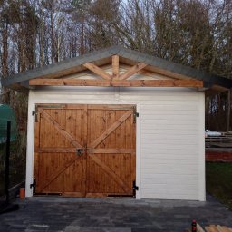 Ogólnobudowlane - Biały budynek gospodarczy z drewnianymi, dwuskrzydłowymi drzwiami i szarym dachem, otoczony kostką brukową. Widoczne elementy konstrukcji drewnianej dachu.