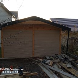 Ogólnobudowlane - Nowo wybudowany, beżowy budynek gospodarczy z drewnianą konstrukcją dachu, widoczny z zewnątrz, z narzędziami i deskami w pobliżu, na tle innych budynków.