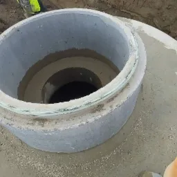 Betonowa studnia kanalizacyjna z uszczelnieniem na styku segmentów, widoczna z góry w wykopie, z fragmentem odzieży roboczej w prawym dolnym rogu.