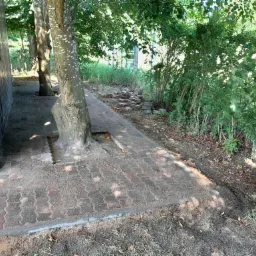 Ułożona kostka brukowa w odcieniach czerwieni i brązu tworząca ścieżkę wokół drzewa, z widocznym obrzeżem i pozostałościami piasku na powierzchni, przylegająca do metalowego garażu i zarośli.