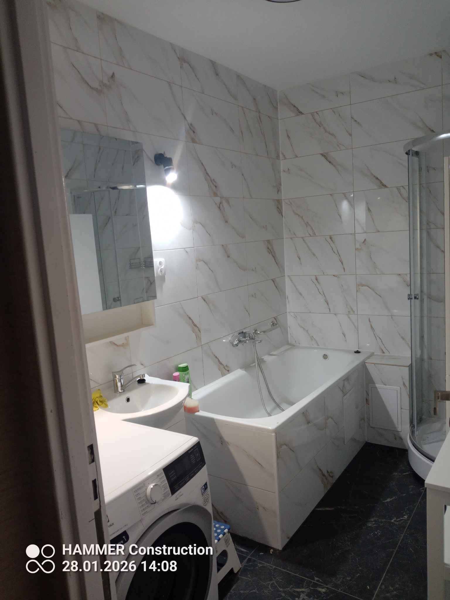 Wykończona łazienka z wanną obudowaną płytkami imitującymi marmur, kabiną prysznicową i pralką. Umywalka z lustrem i czarna podłoga.