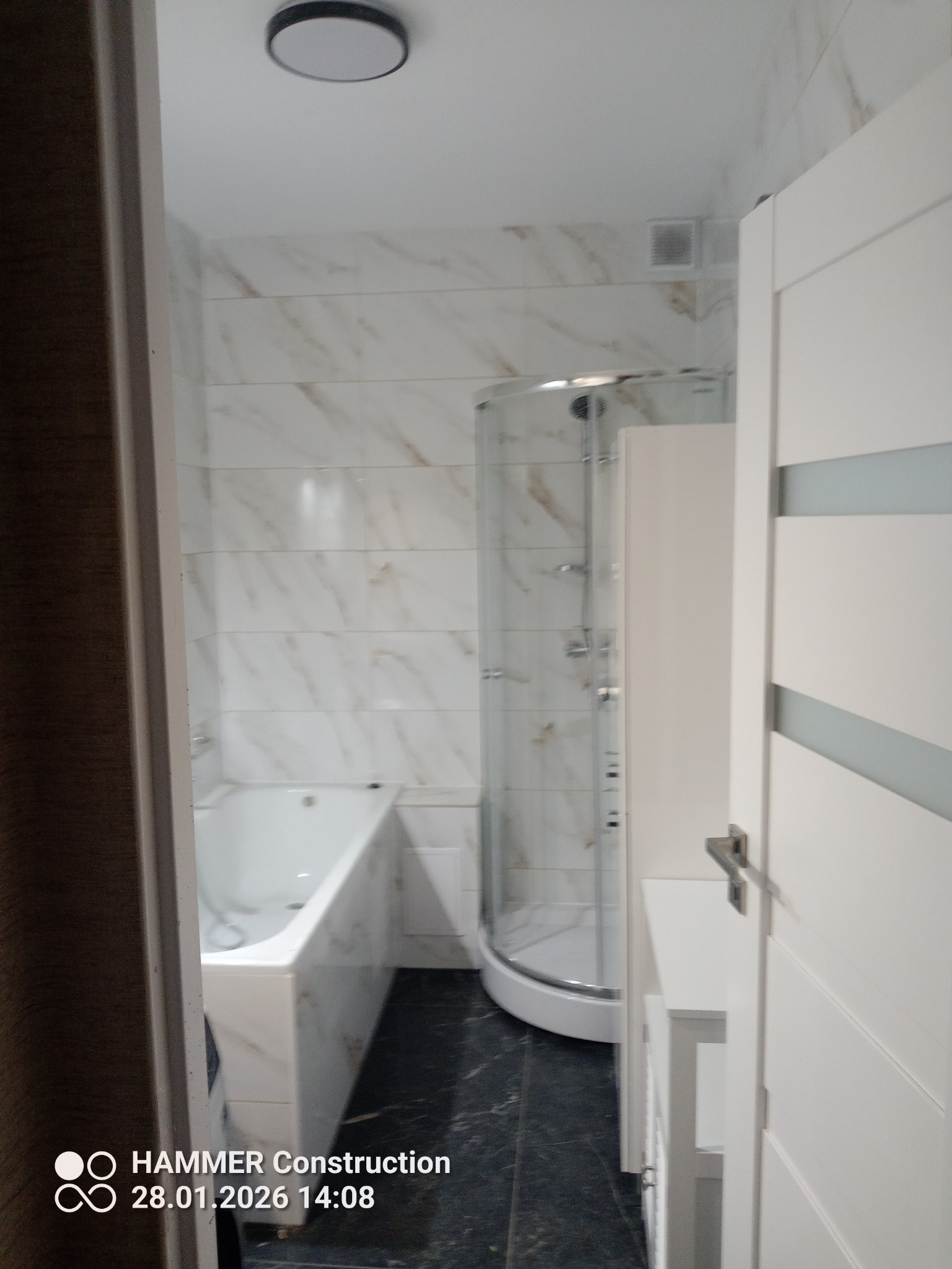 Nowoczesna łazienka po remoncie: białe marmurowe płytki, kabina prysznicowa z zaokrąglonym szkłem, wanna obudowana płytkami i czarna podłoga. Widok zza uchylonych drzwi.