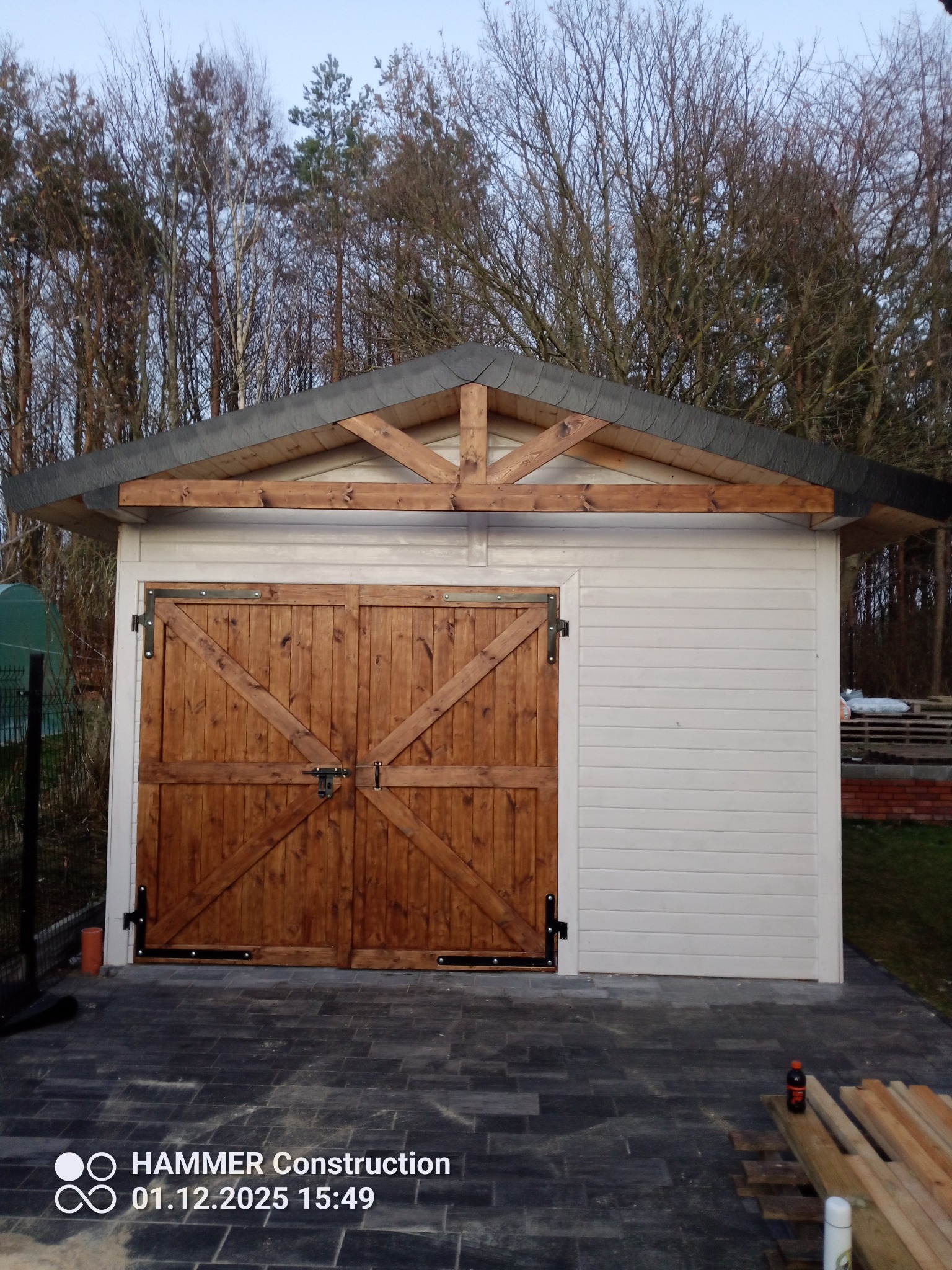 Biały budynek gospodarczy z drewnianymi, dwuskrzydłowymi drzwiami i szarym dachem, otoczony kostką brukową. Widoczne elementy konstrukcji drewnianej dachu.