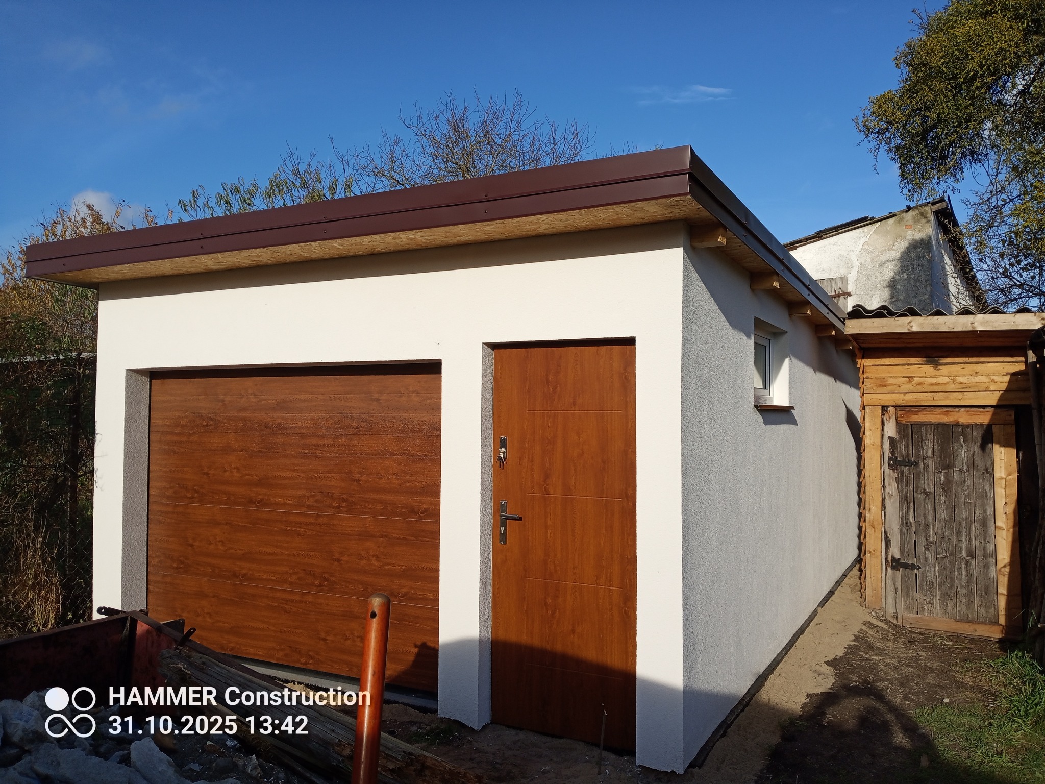Nowo wybudowany, parterowy garaż z brązową bramą segmentową i drzwiami wejściowymi, wykończony jasnym tynkiem zewnętrznym. Widoczne elementy dachu i otoczenie.