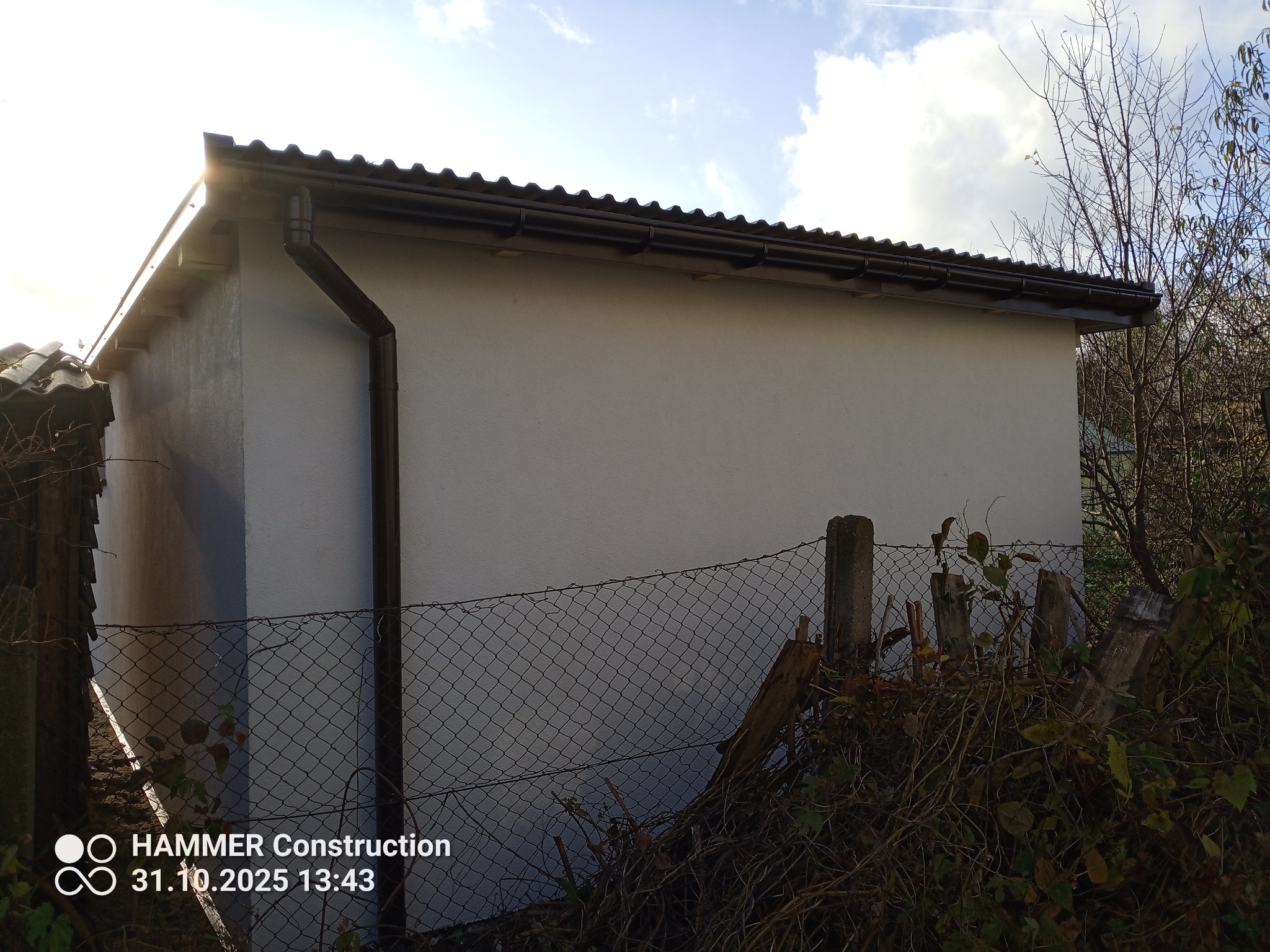 Nowy, otynkowany budynek gospodarczy z brązowymi rynnami i dachem z blachy trapezowej, widziany zza ogrodzenia z siatki i zarośli.