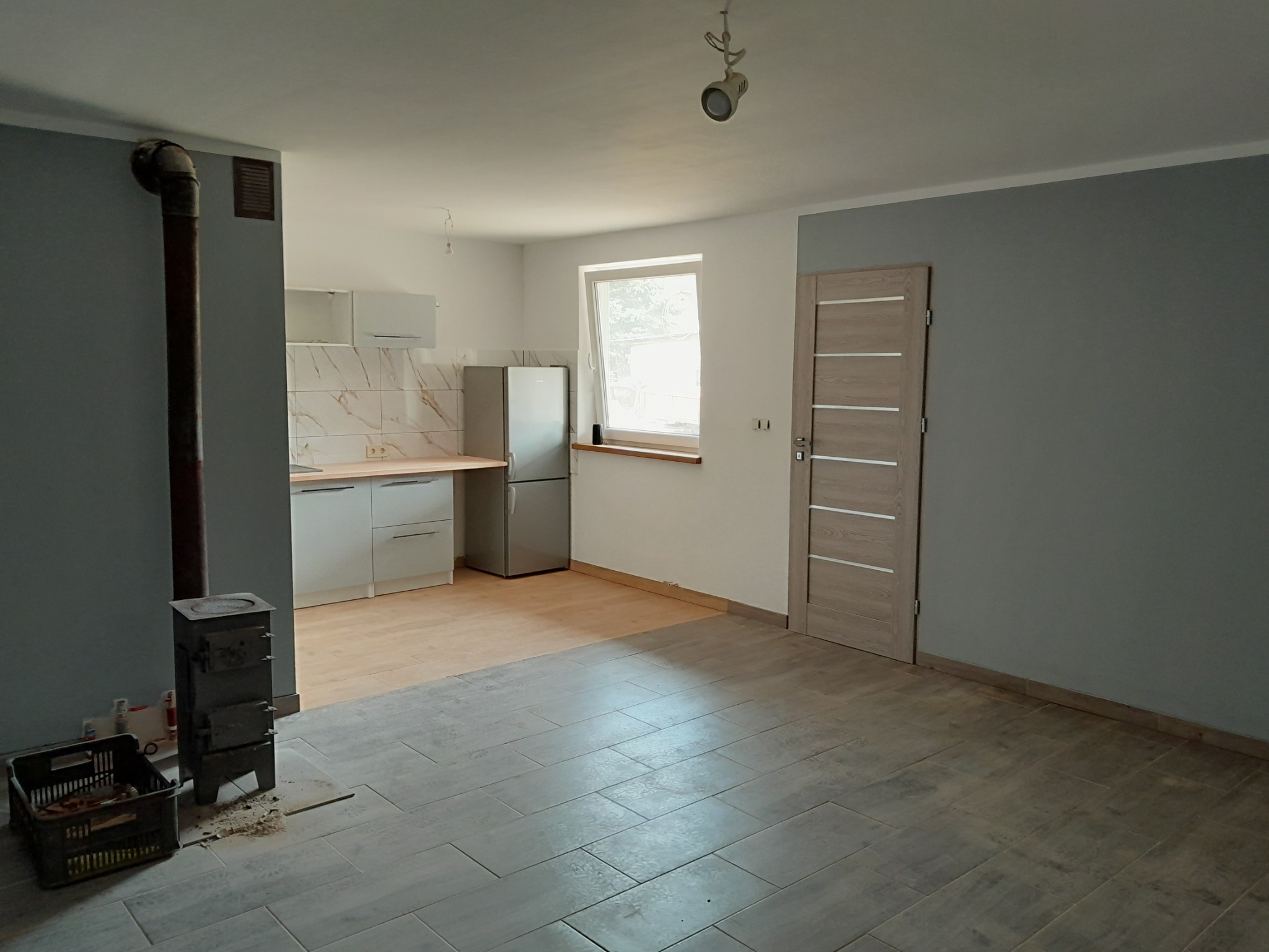 Wnętrze po remoncie: szare ściany, drewnopodobne płytki podłogowe, kuchnia z białymi szafkami i srebrną lodówką, piec wolnostojący z rurą dymową, drzwi wewnętrzne z poziomymi panelami.