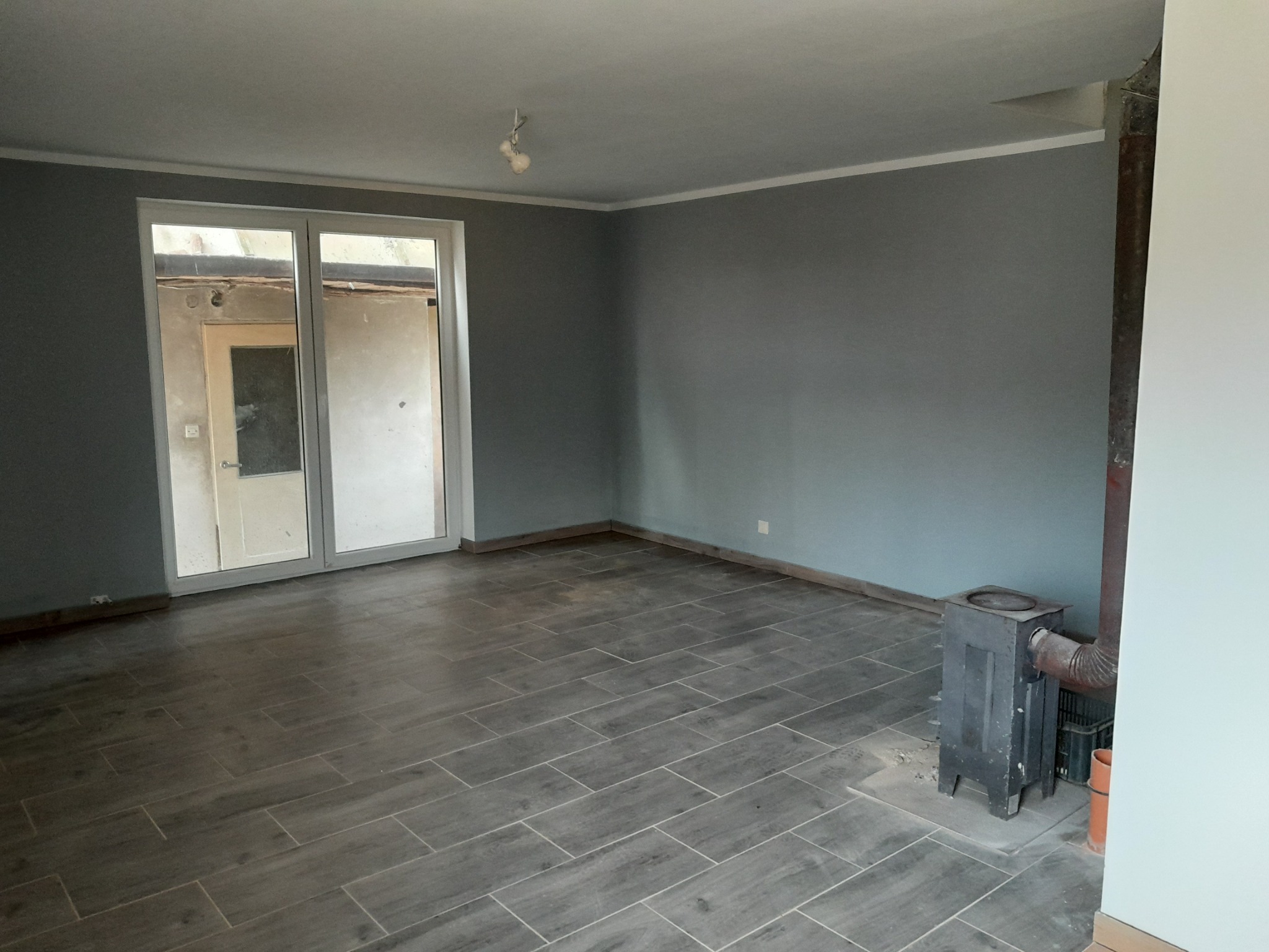 Szare, wykafelkowane pomieszczenie z piecem wolnostojącym i widokiem przez okno na sąsiedni budynek w trakcie prac remontowych.