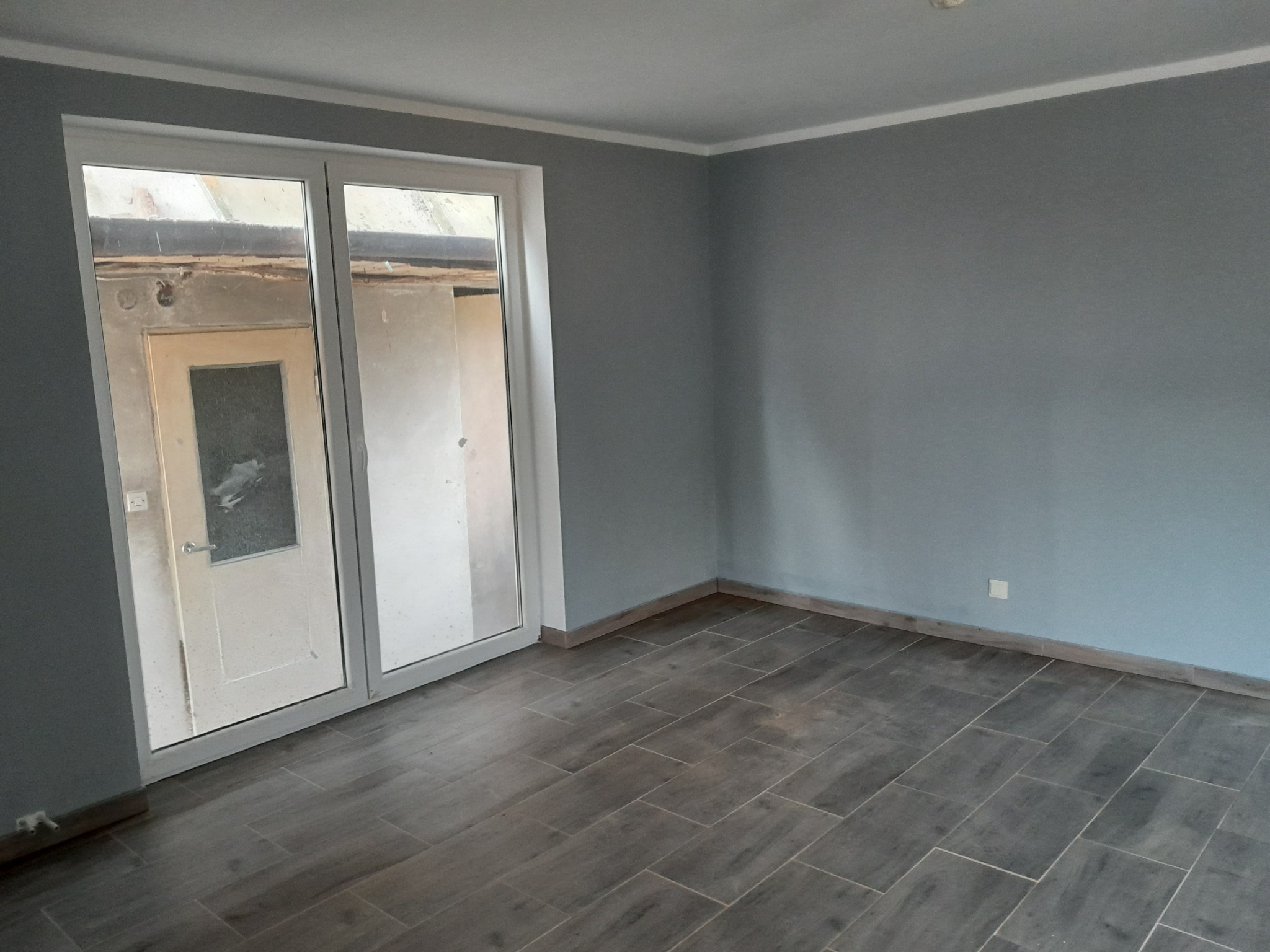 Szare płytki podłogowe imitujące drewno w pustym pokoju z szarymi ścianami i białymi listwami przypodłogowymi oraz białymi drzwiami balkonowymi.