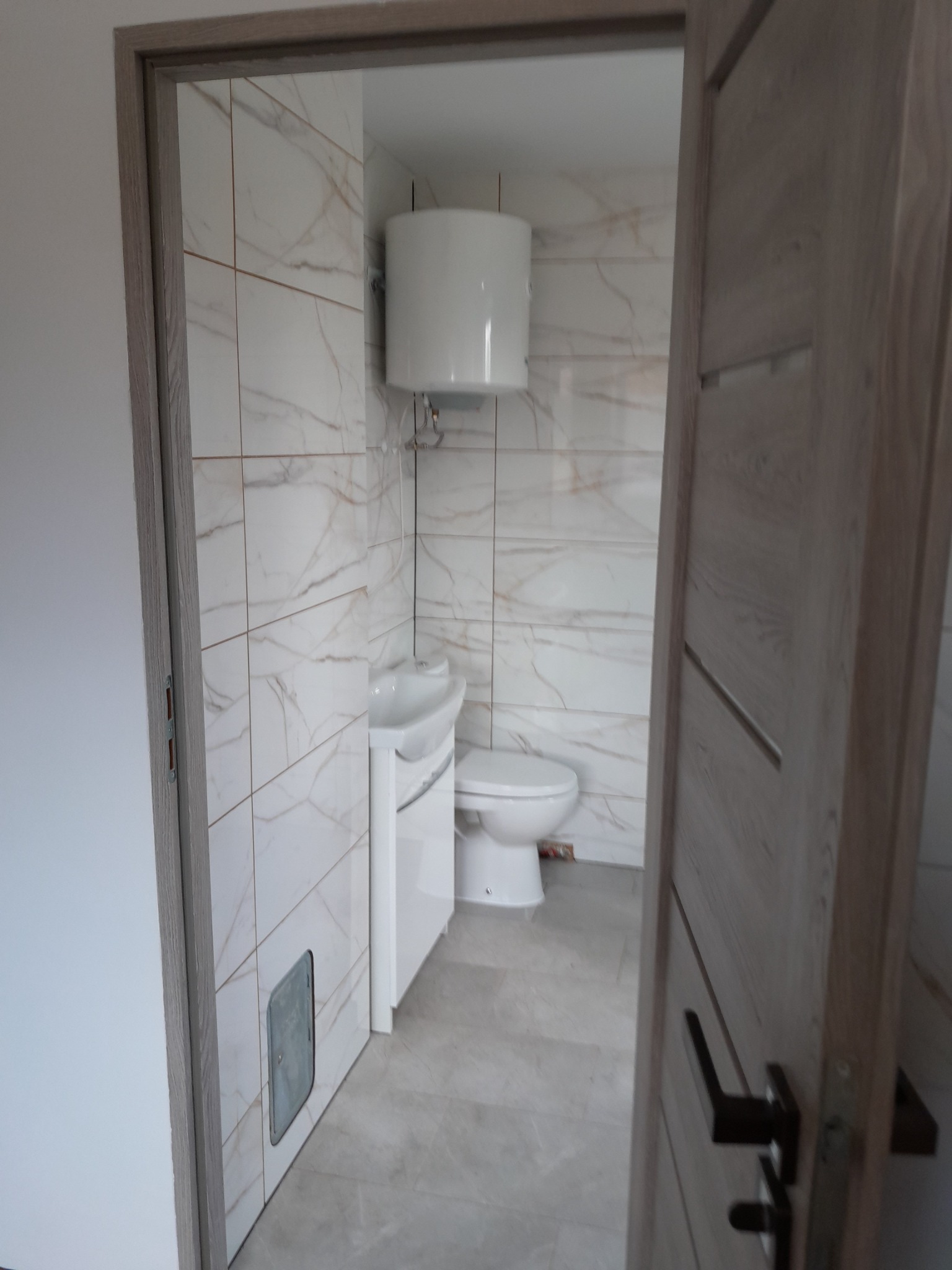 Wnętrze wykończonej łazienki z toaletą, umywalką i bojlerem, wyłożonej płytkami imitującymi marmur, widoczne przez otwarte drzwi z drewnianą ościeżnicą.