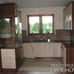 Nowoczesna kuchnia z beżowymi frontami szafek i ciemnobrązowymi elementami drewnianymi, przeszklona witryna z szufladami po lewej stronie, okno z widokiem na zieleń.