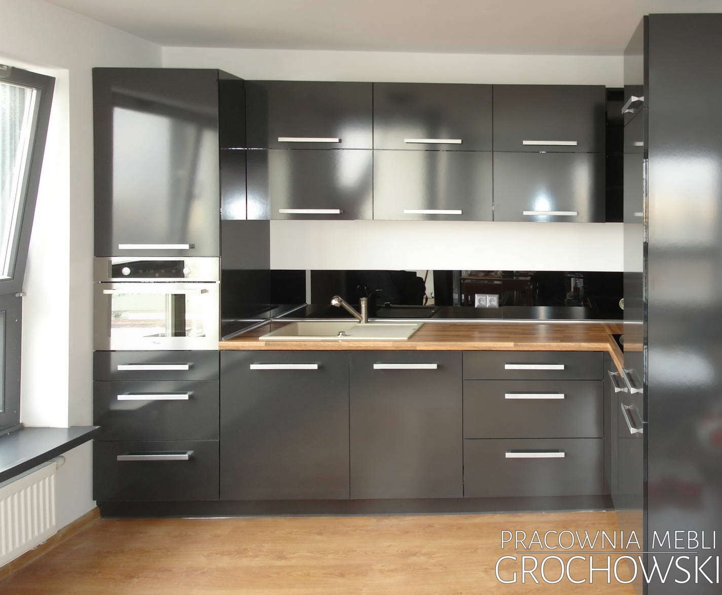 Nowoczesna kuchnia z czarnymi, lakierowanymi frontami, drewnianym blatem i minimalistycznymi, srebrnymi uchwytami, oświetlona naturalnym światłem wpadającym przez okno.