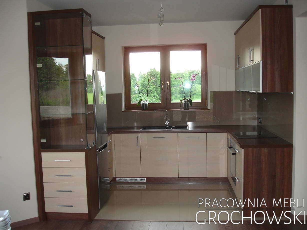 Nowoczesna kuchnia z beżowymi frontami szafek i ciemnobrązowymi elementami drewnianymi, przeszklona witryna z szufladami po lewej stronie, okno z widokiem na zieleń.