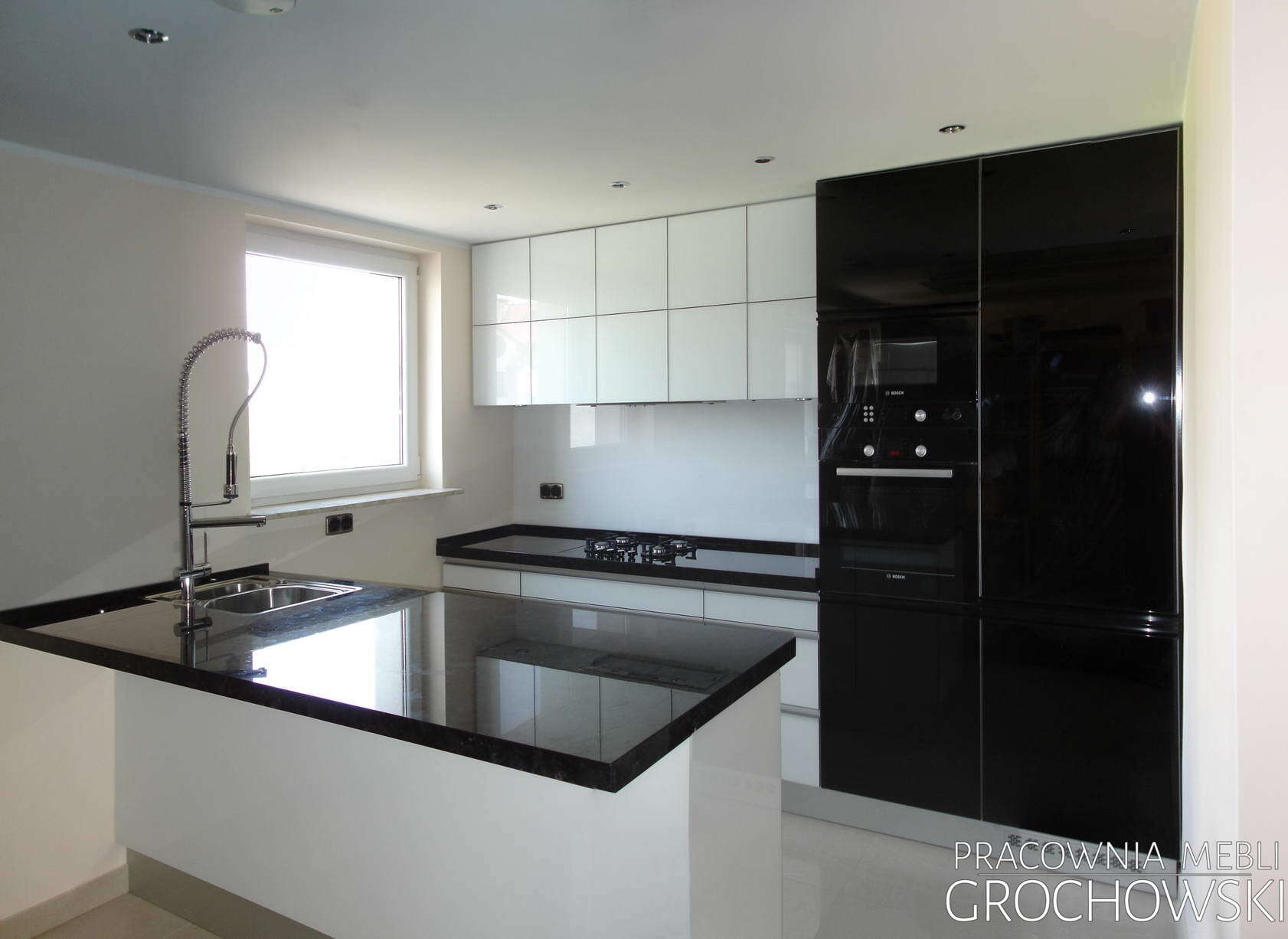 Nowoczesna kuchnia z czarnym blatem, białymi szafkami wiszącymi, czarną zabudową piekarnika i wyspą kuchenną z chromowaną baterią.