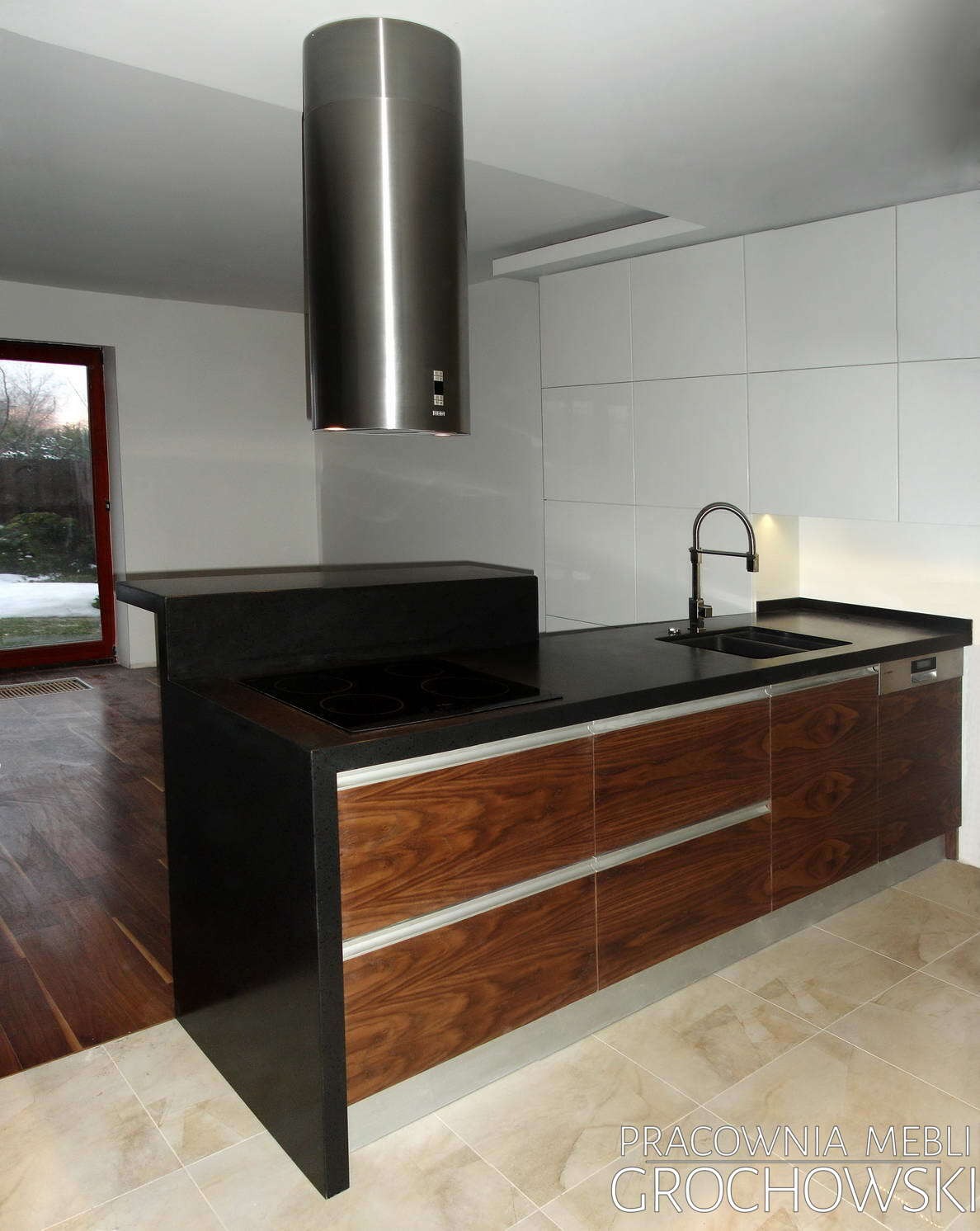 Nowoczesna kuchnia z wyspą: ciemny blat, drewniane fronty szafek z metalowymi uchwytami, cylindryczny okap ze stali nierdzewnej. Białe, gładkie szafki wiszące w tle.