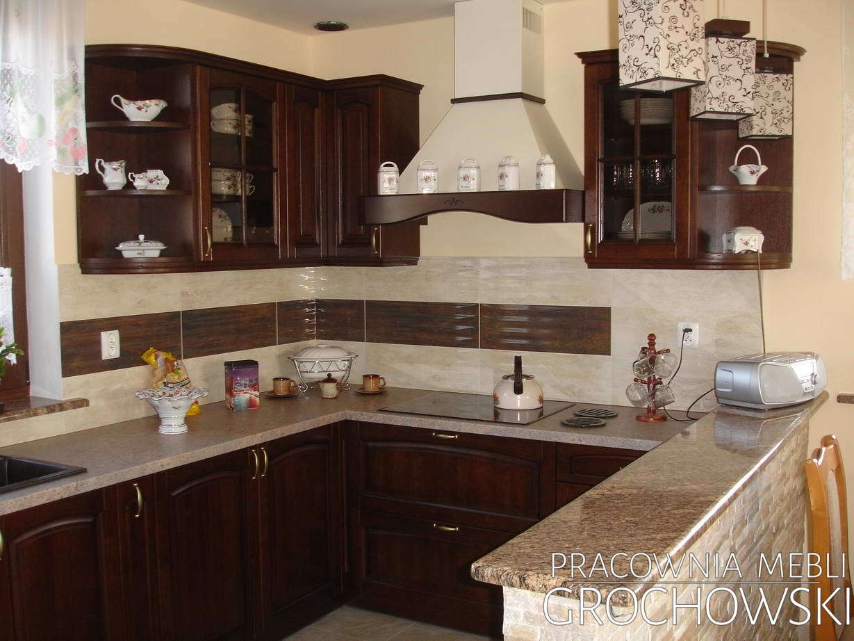 Klasyczna kuchnia z ciemnego drewna z przeszklonymi szafkami wiszącymi, wyposażona w płytę indukcyjną i okap, z blatem wykończonym w jasnym odcieniu, dekoracje na blacie i lampa z abażurem...