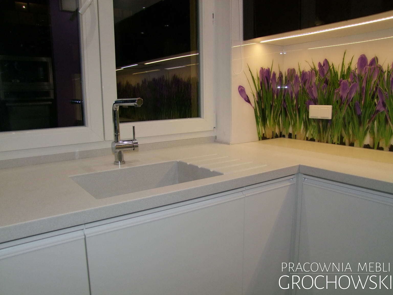 Jasna, minimalistyczna kuchnia z blatem z konglomeratu, chromowaną baterią i panelem ściennym z motywem fioletowych krokusów podświetlonym ledami.