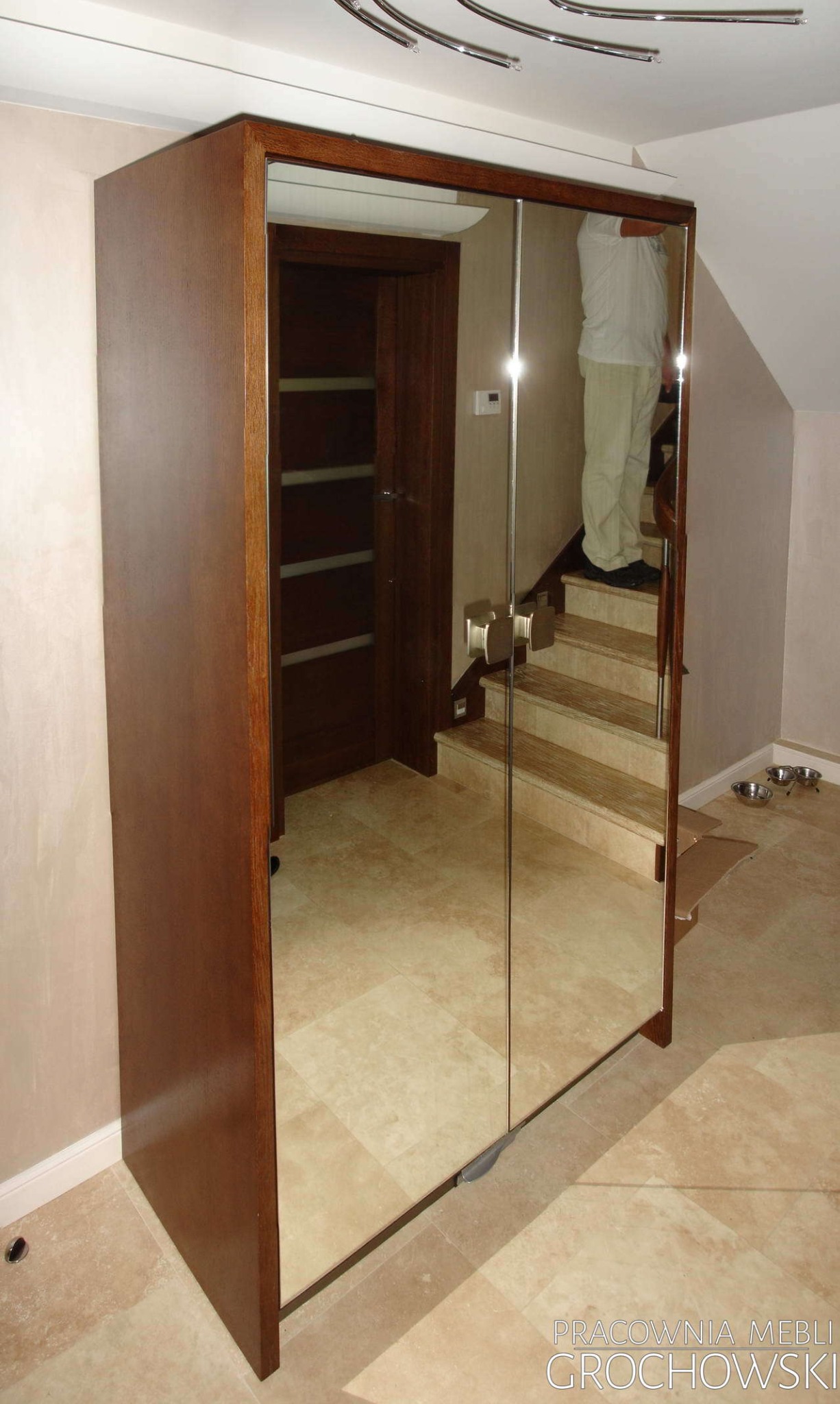 Wysoka, brązowa szafa z przesuwnymi drzwiami lustrzanymi, odbijająca fragment schodów i drzwi w tle.