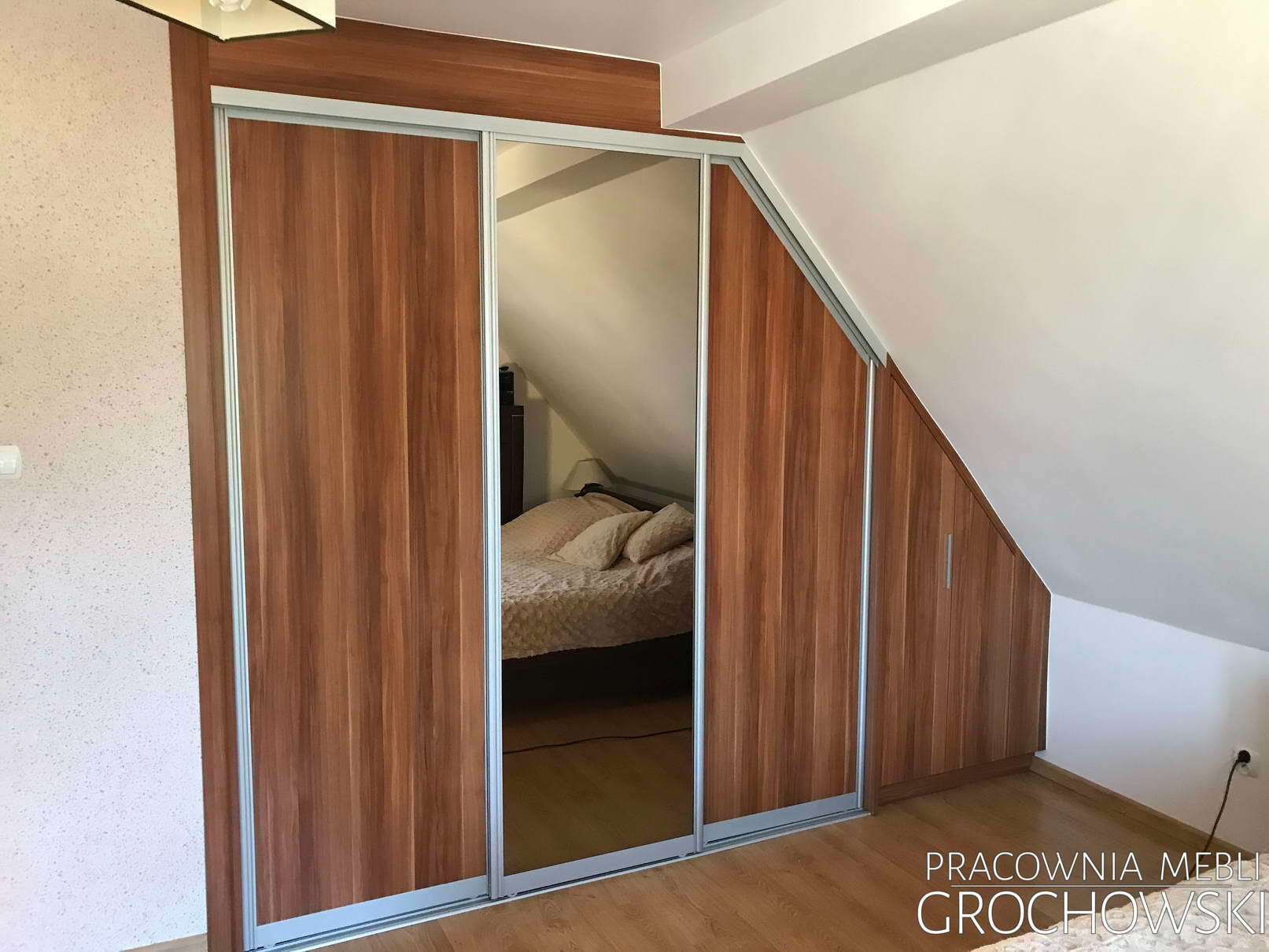 Szafa wnękowa z przesuwnymi drzwiami z lustrem, dopasowana do skosu poddasza, z widocznym odbiciem łóżka i szafki nocnej w lustrze.