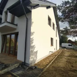 Świeżo otynkowana ściana domu jednorodzinnego z grafitową rynną spustową, widoczne okna z brązowymi ramami, obok ułożona kostka brukowa i przygotowane podłoże pod chodnik, w tle biały bus.