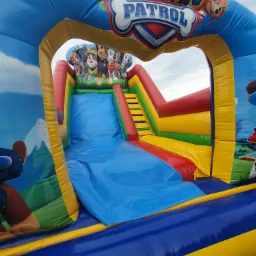 Kolorowy dmuchany zamek ze zjeżdżalnią Paw Patrol, widok z dołu, na zielonej trawie.