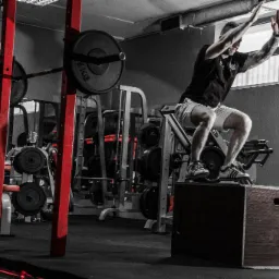 Mężczyzna z tatuażami w sportowym stroju w dynamicznym skoku z drewnianej skrzyni w siłowni, w tle maszyny do ćwiczeń i sztangi.